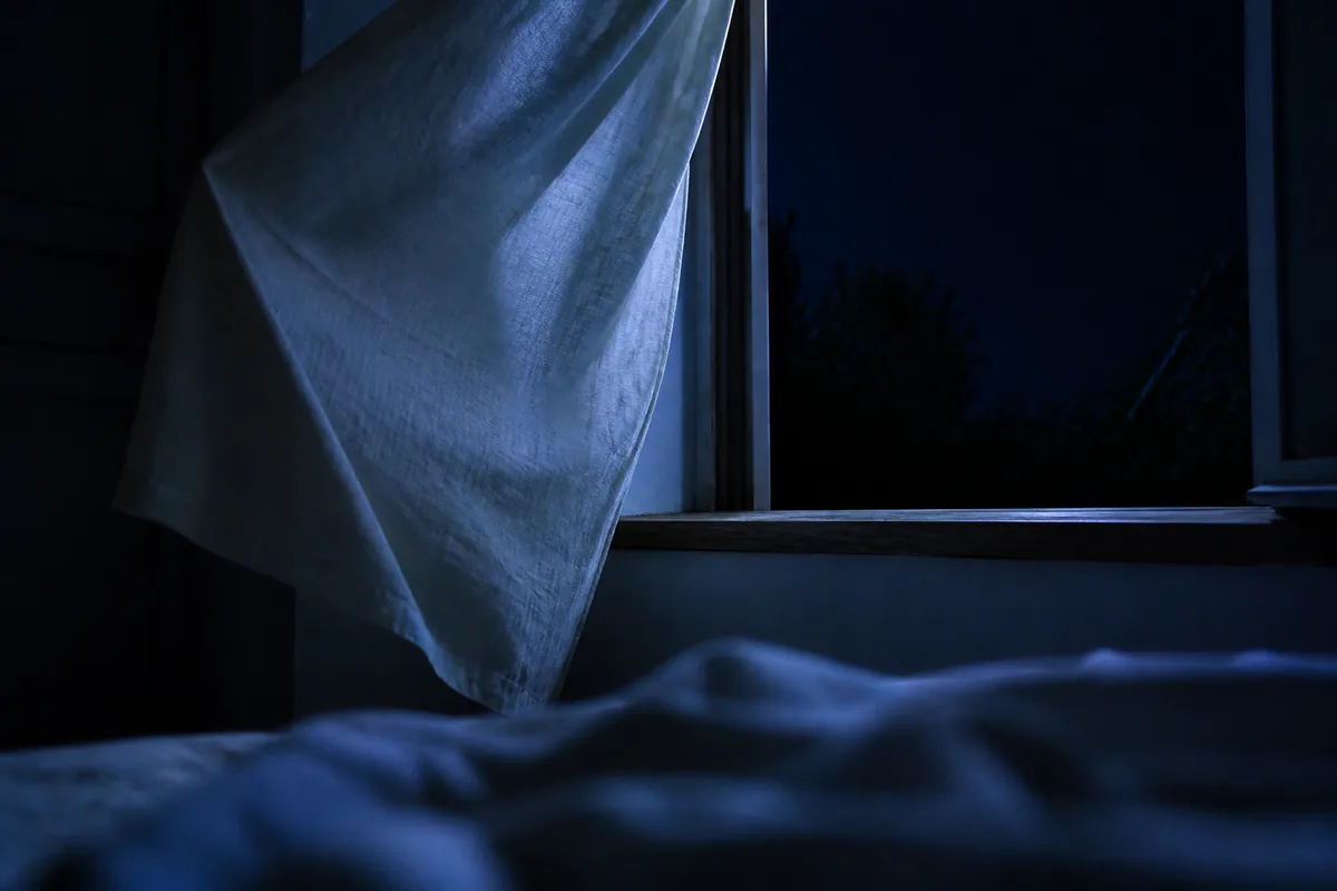 Fenêtre ouverte la nuit sur une chambre sombre, voilage de lin soulevé par un courant d’air