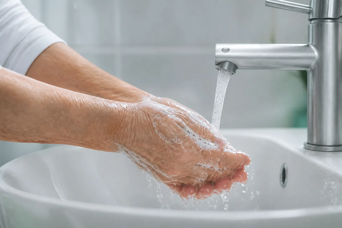 Mains mûres couvertes de mousse rincées sous un filet d&rsquo;eau tiède au-dessus d&rsquo;une vasque en céramique blanche